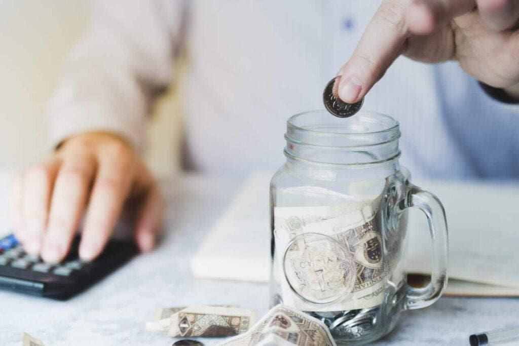 Person using a calculator next to a jar of coins and banknotes, representing cost savings and operational efficiency in equipment rentals through asset tracking