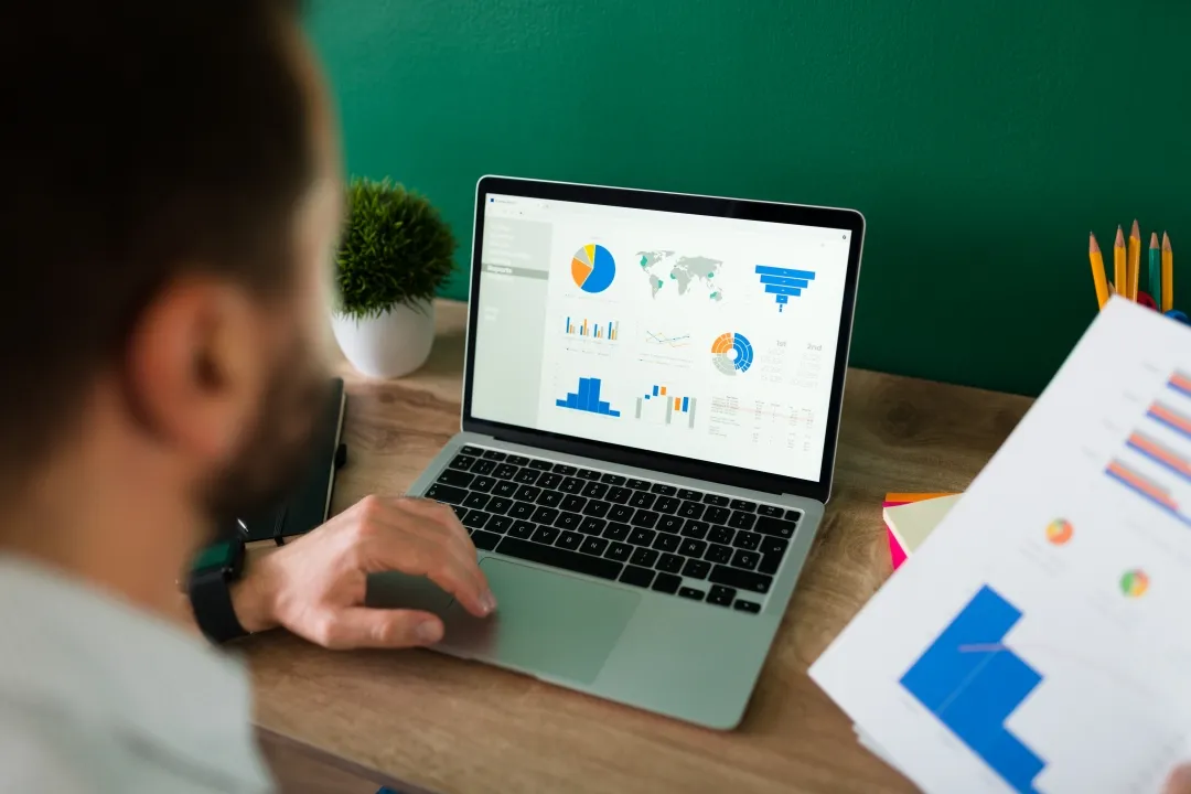 A man focused on his laptop, surrounded by graphs and data visualizations on the desk.