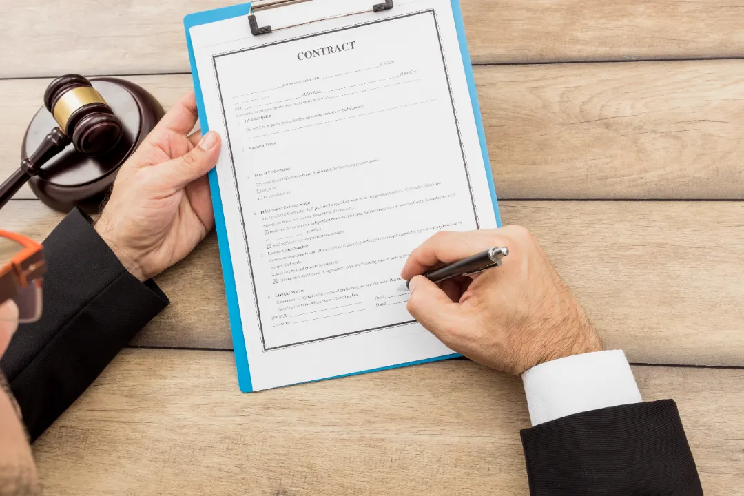 Person signing a contract on a clipboard with a gavel on the table.
