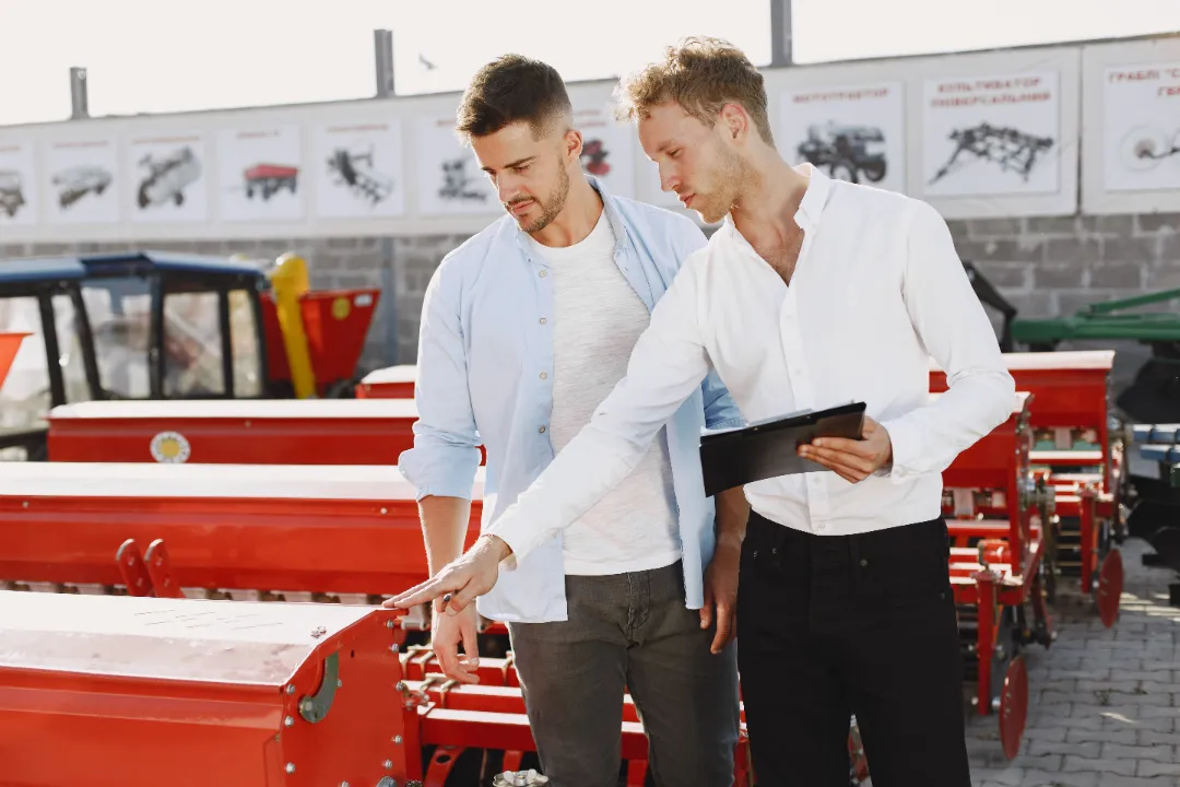 Two people discussing farming equipment with a tablet in front of a red machine.